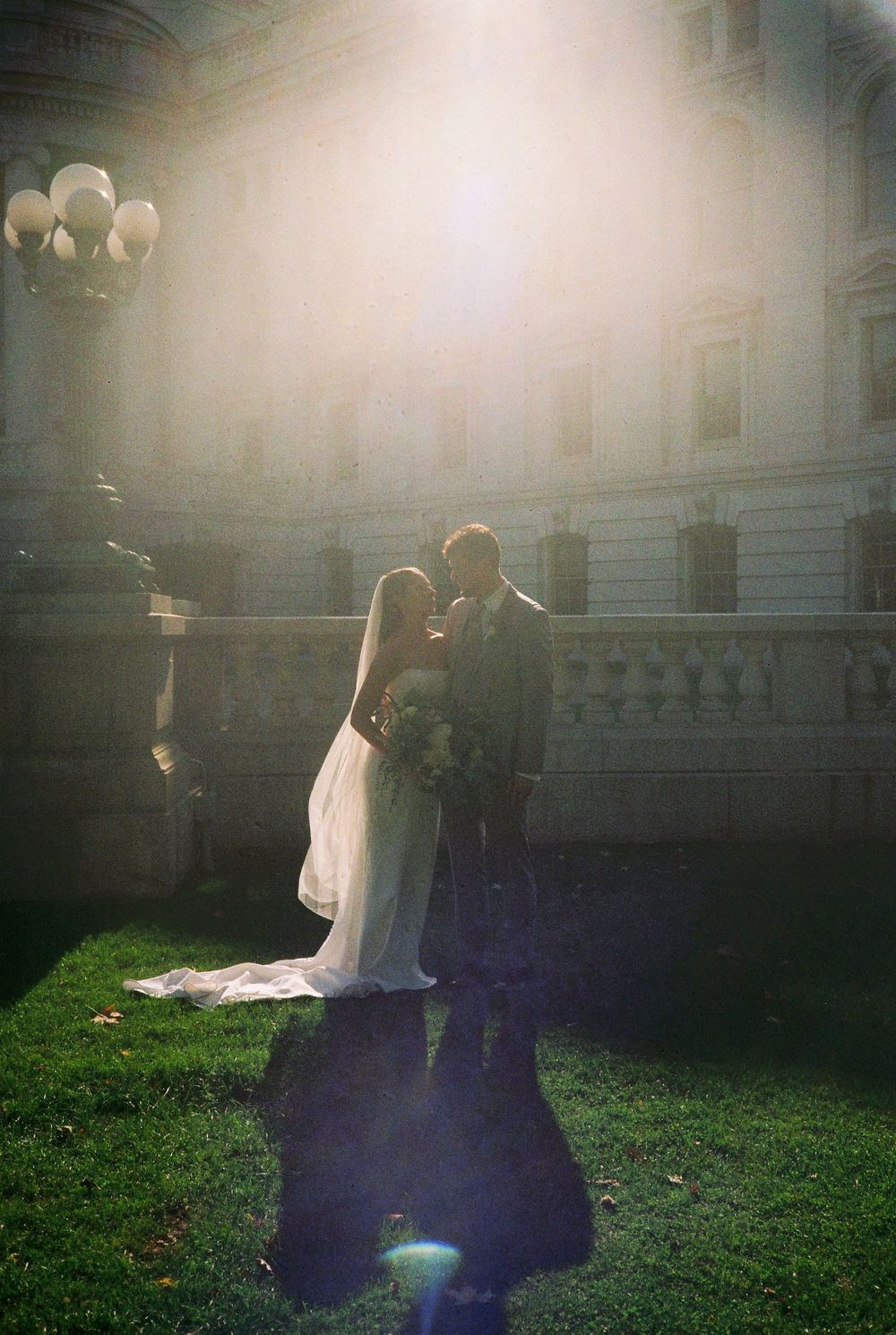 A couple in wedding attire embraces on a grassy lawn at sunset with a glowing lens flare effect.