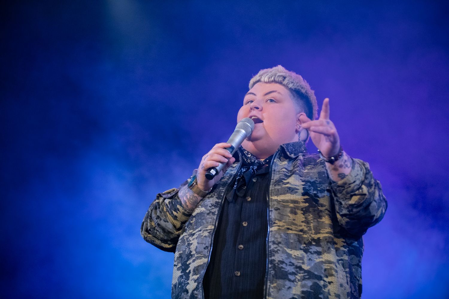 Singer performing on stage with microphone in colorful blue smoke at live concert event.