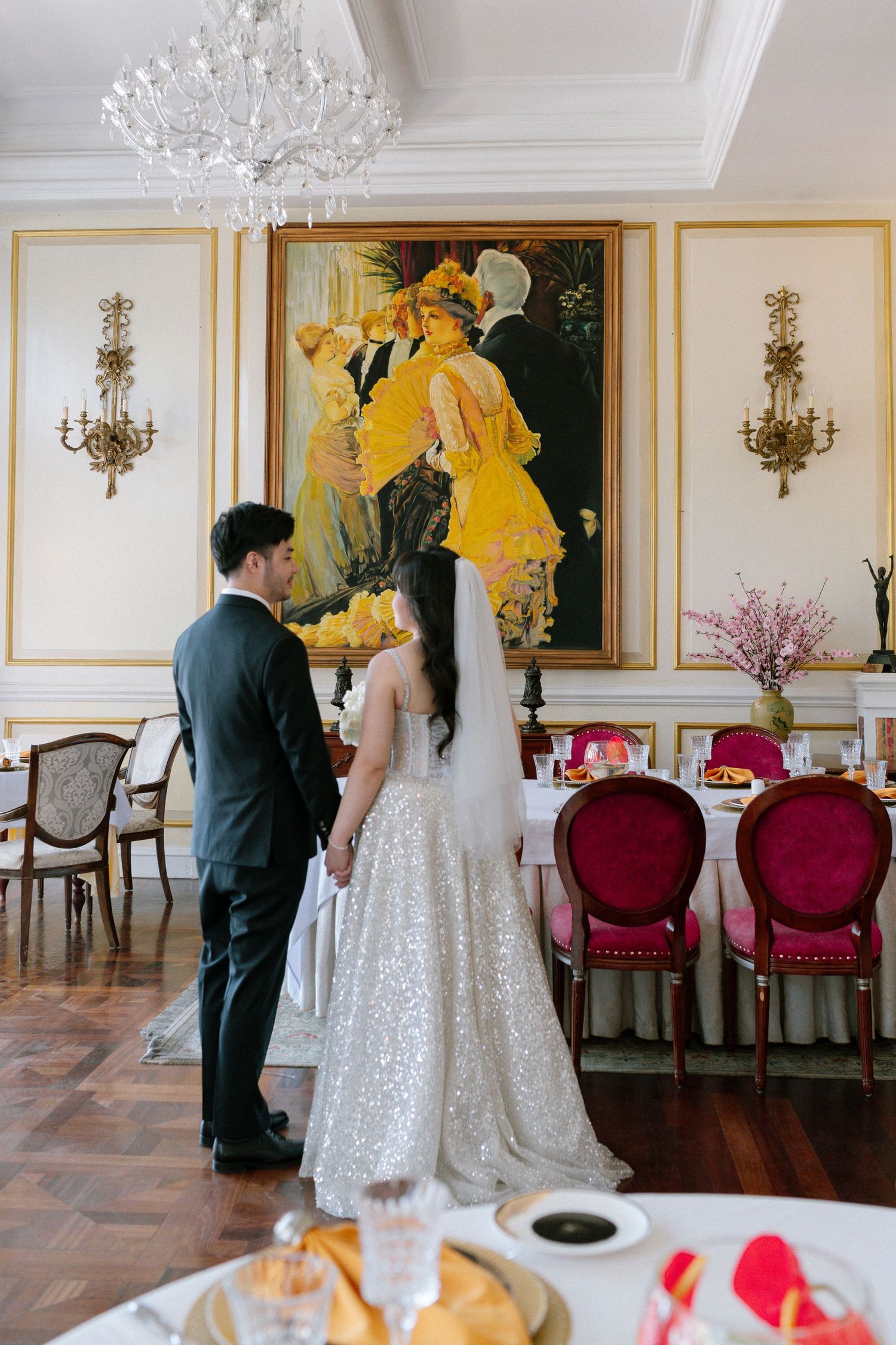 Bride and groom hold hands admiring elegant dining room with ornate painting and crystal chandelier.
