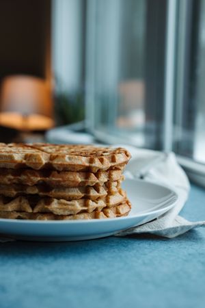 Close-up side view of a stack of golden homemade waffles on a white plate by a window.