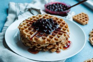 Stack of golden waffles topped with blueberry compote on a white plate with jam jar in background.