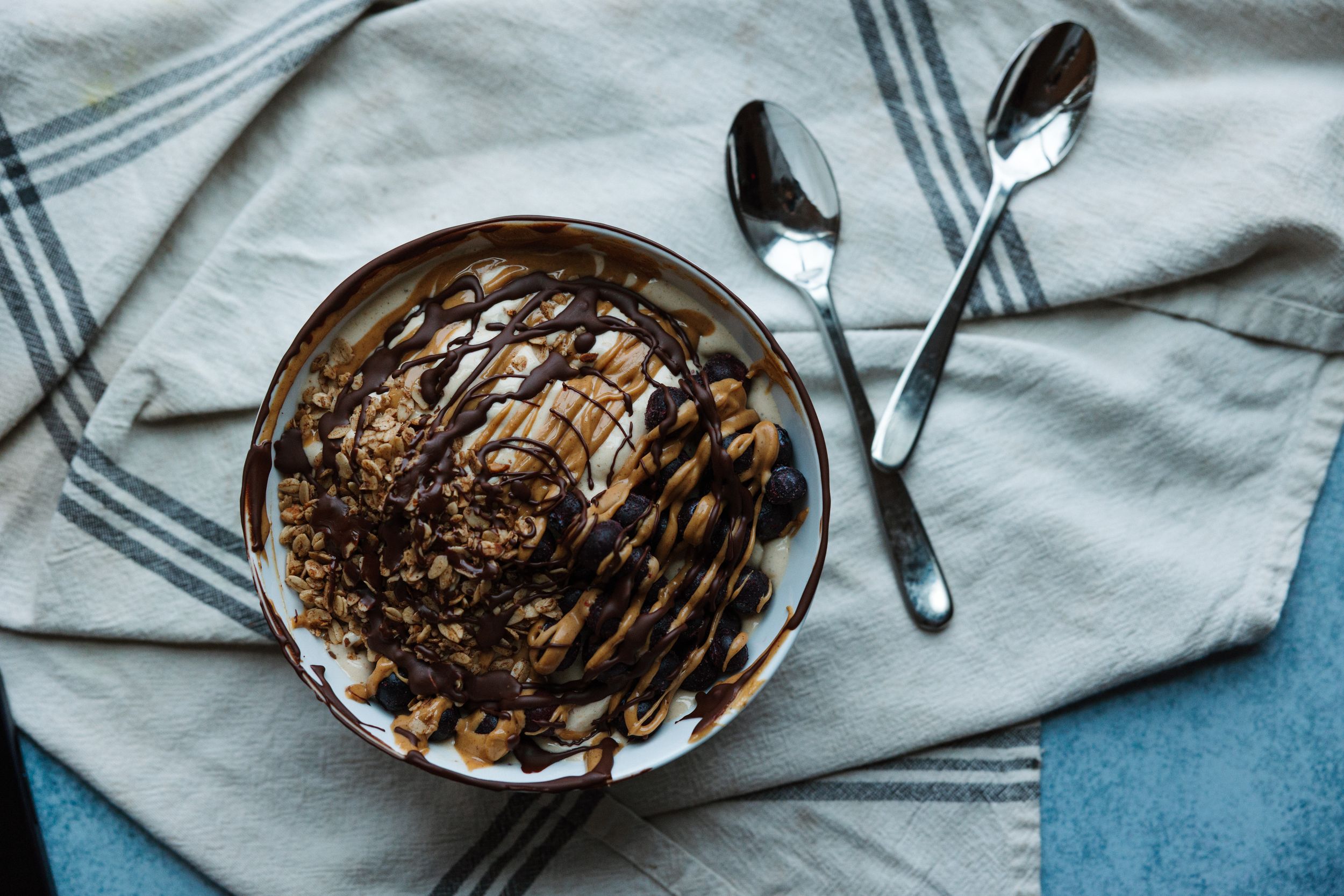 Overhead view of a bowl of granola with peanut butter and chocolate drizzle on striped cloth.