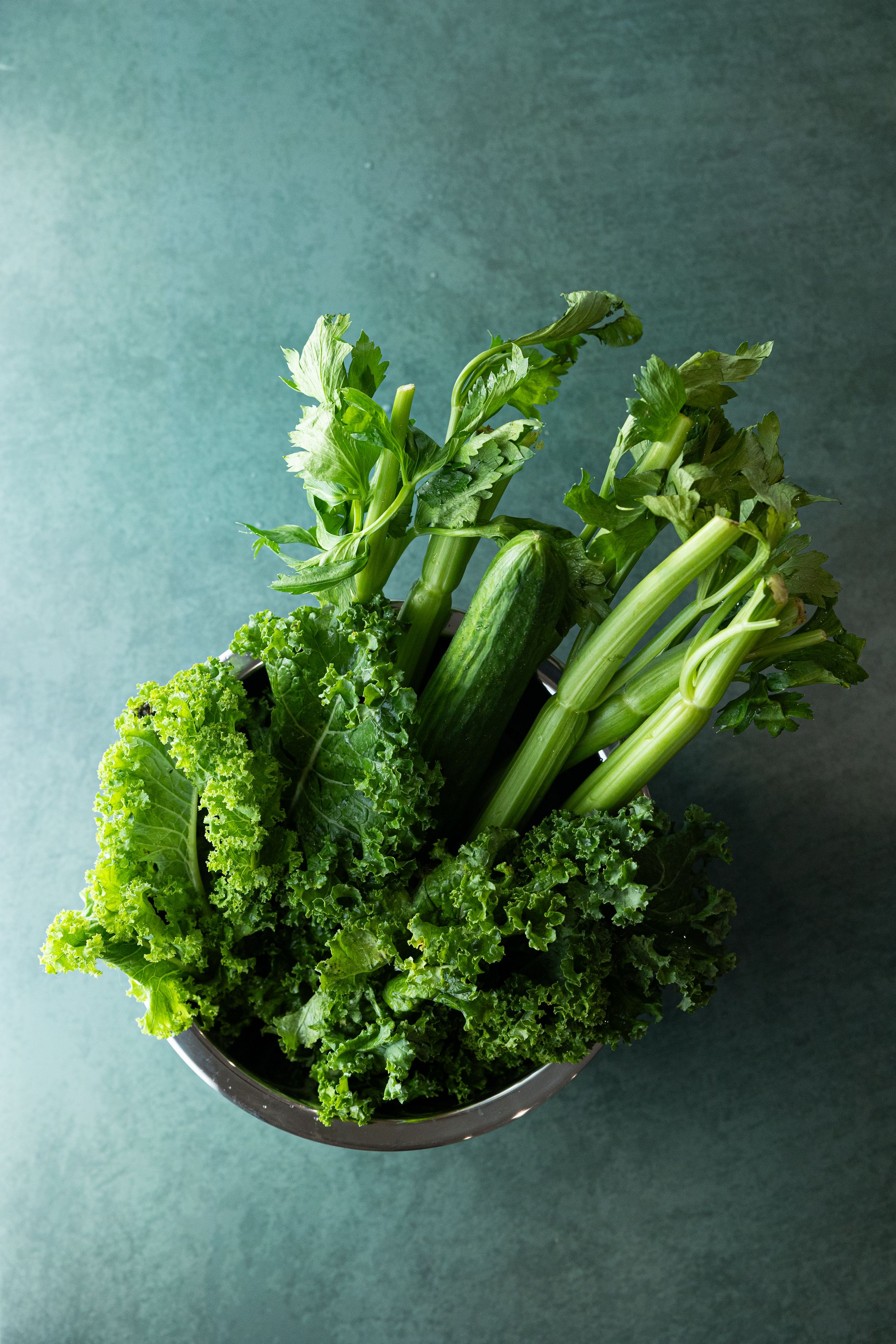 Fresh green vegetables including kale, celery, and cucumber in a colander on dark background.