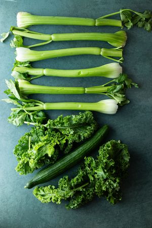 Fresh green celery stalks, kale, and cucumbers arranged on a dark surface.