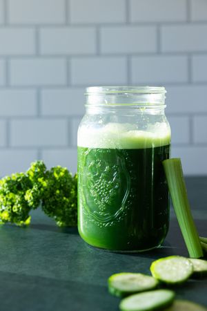 Green juice in a mason jar with kale, celery, and cucumber slices on a dark surface.