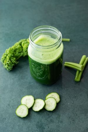 Green juice smoothie in mason jar with fresh kale, celery, and sliced cucumber on dark surface.