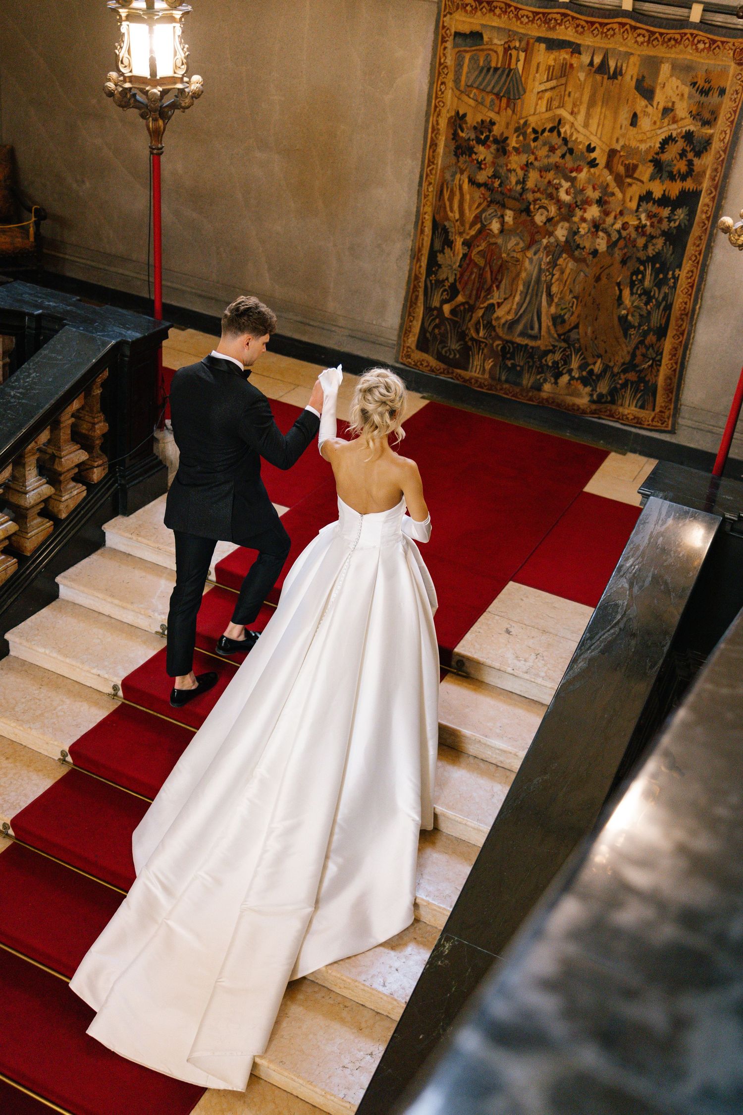 A couple walks down an outdoor aisle lined with flowers and cypress trees leading to an elegant villa entrance.