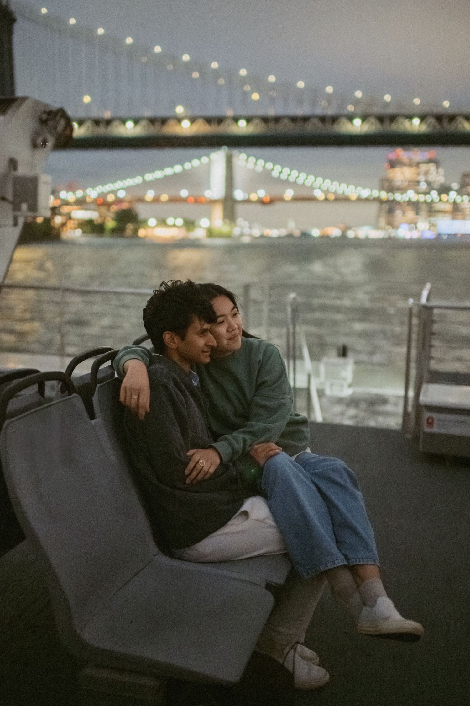 Couple sitting close together on a Brooklyn ferry at dusk, with a lit bridge and city skyline in the background.