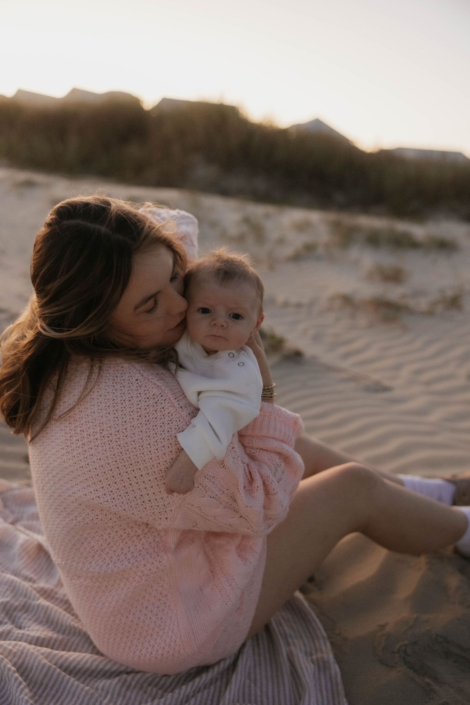Tender moment of a mother in pink knit dress kissing her newborn baby on a sandy beach at golden hour.