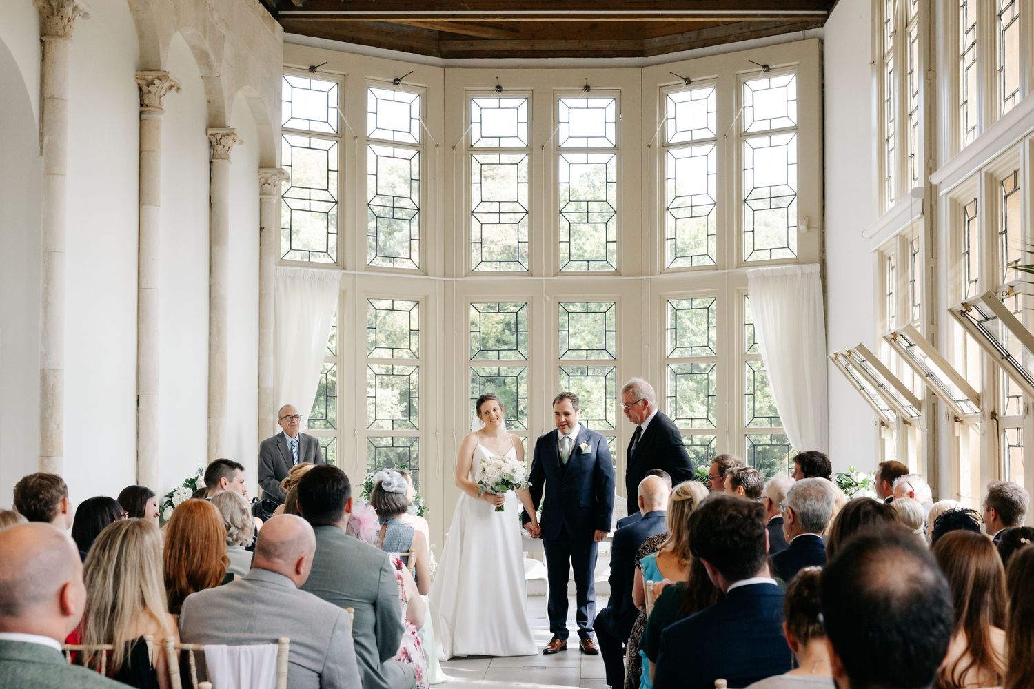 Wedding guests mingle in an elegant white room with large bay windows and ornate architectural details.
