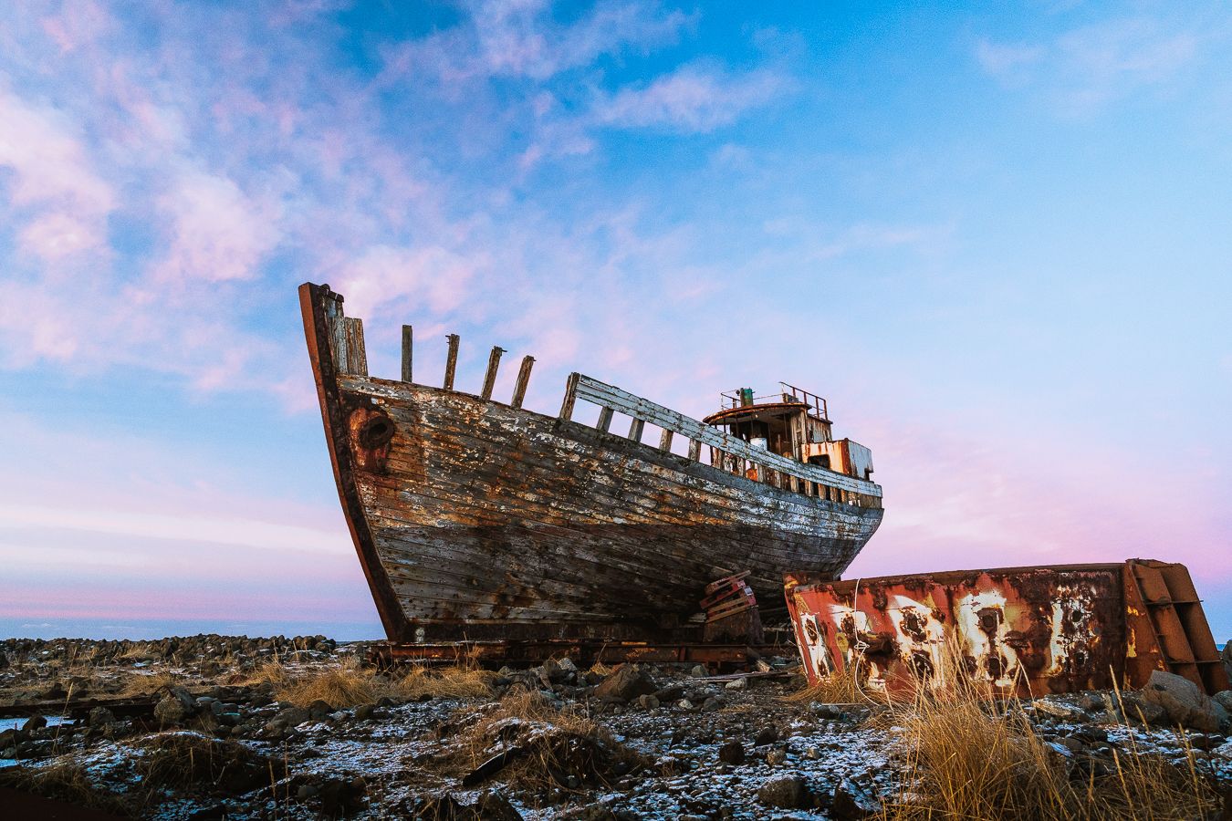 Abandoned wooden shipwreck on rocky shore under dramatic pink and blue sunset sky.