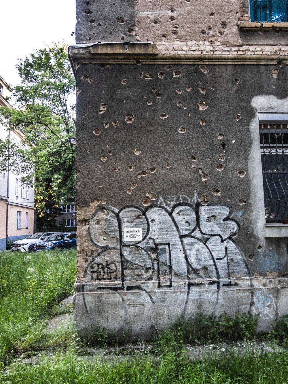 Graffiti tags on bullet-scarred building corner with overgrown grass in urban setting.