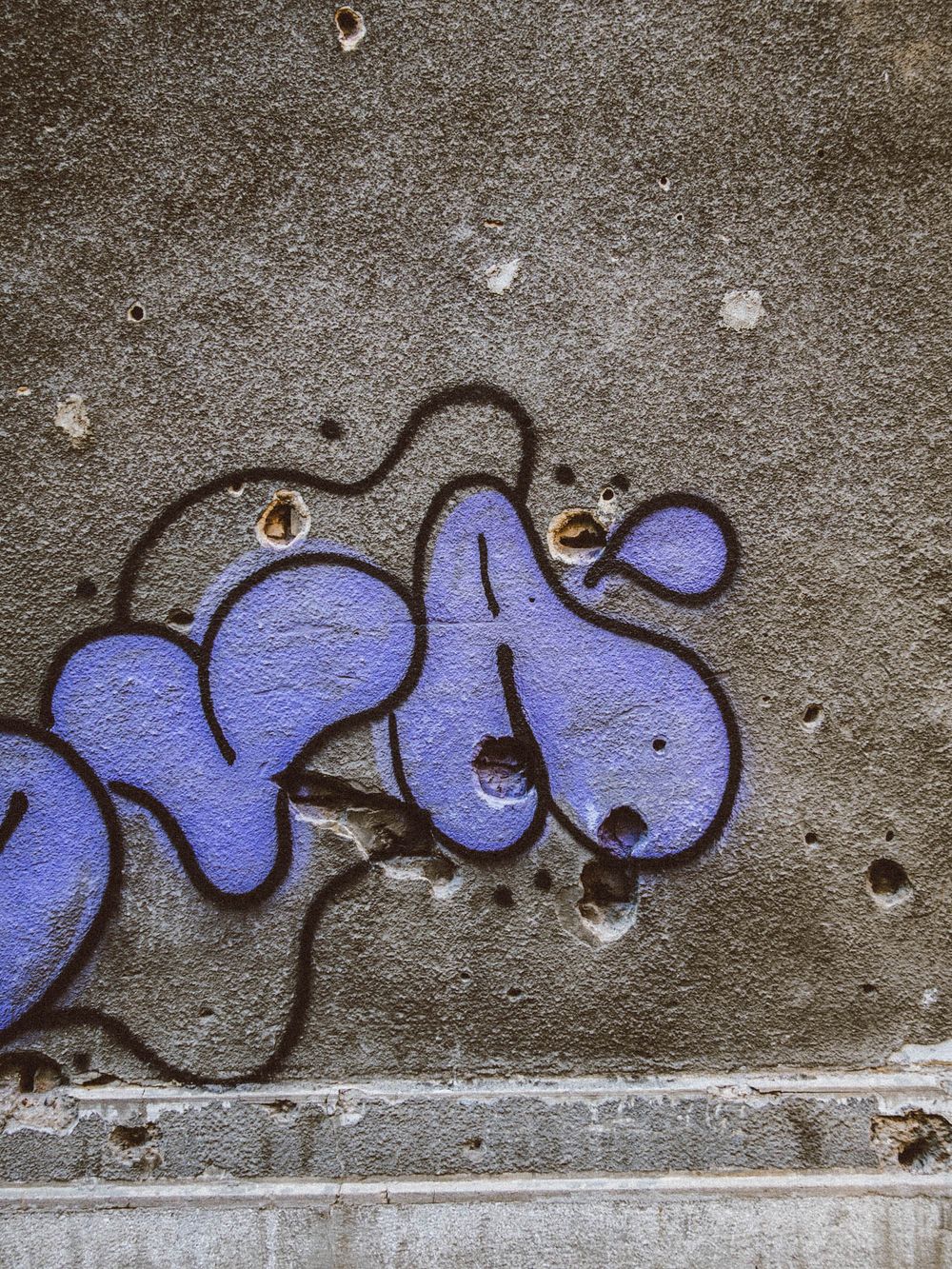 Purple bubble graffiti tag with dark outline on weathered concrete wall with small holes.