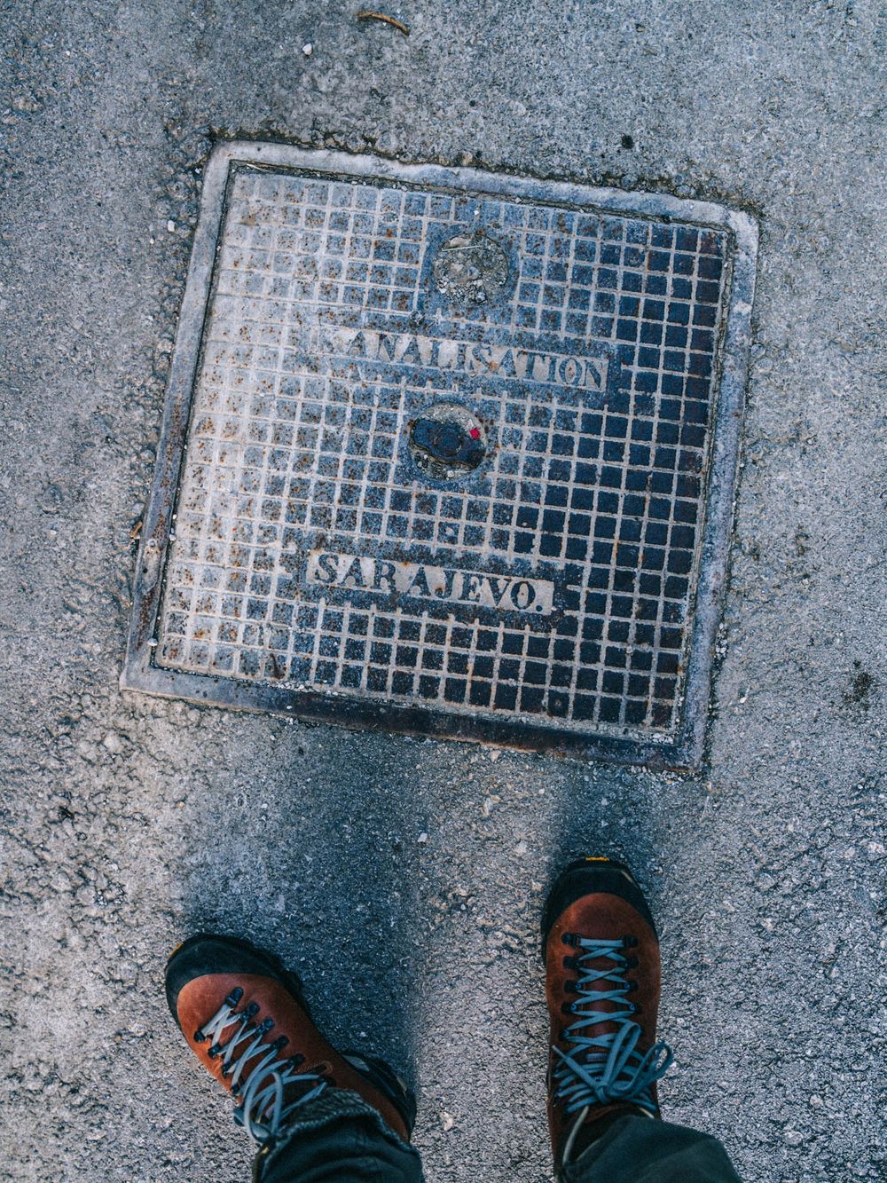 Person standing next to a Sarajevo sanalisation manhole cover on a concrete street.