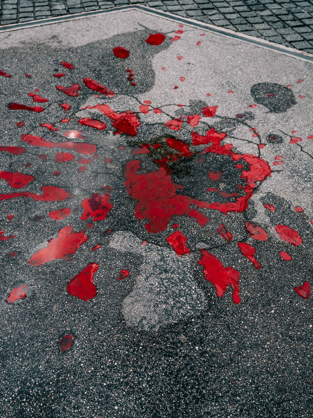 Red liquid splattered on gray asphalt pavement, creating a dramatic abstract pattern near a tiled sidewalk.