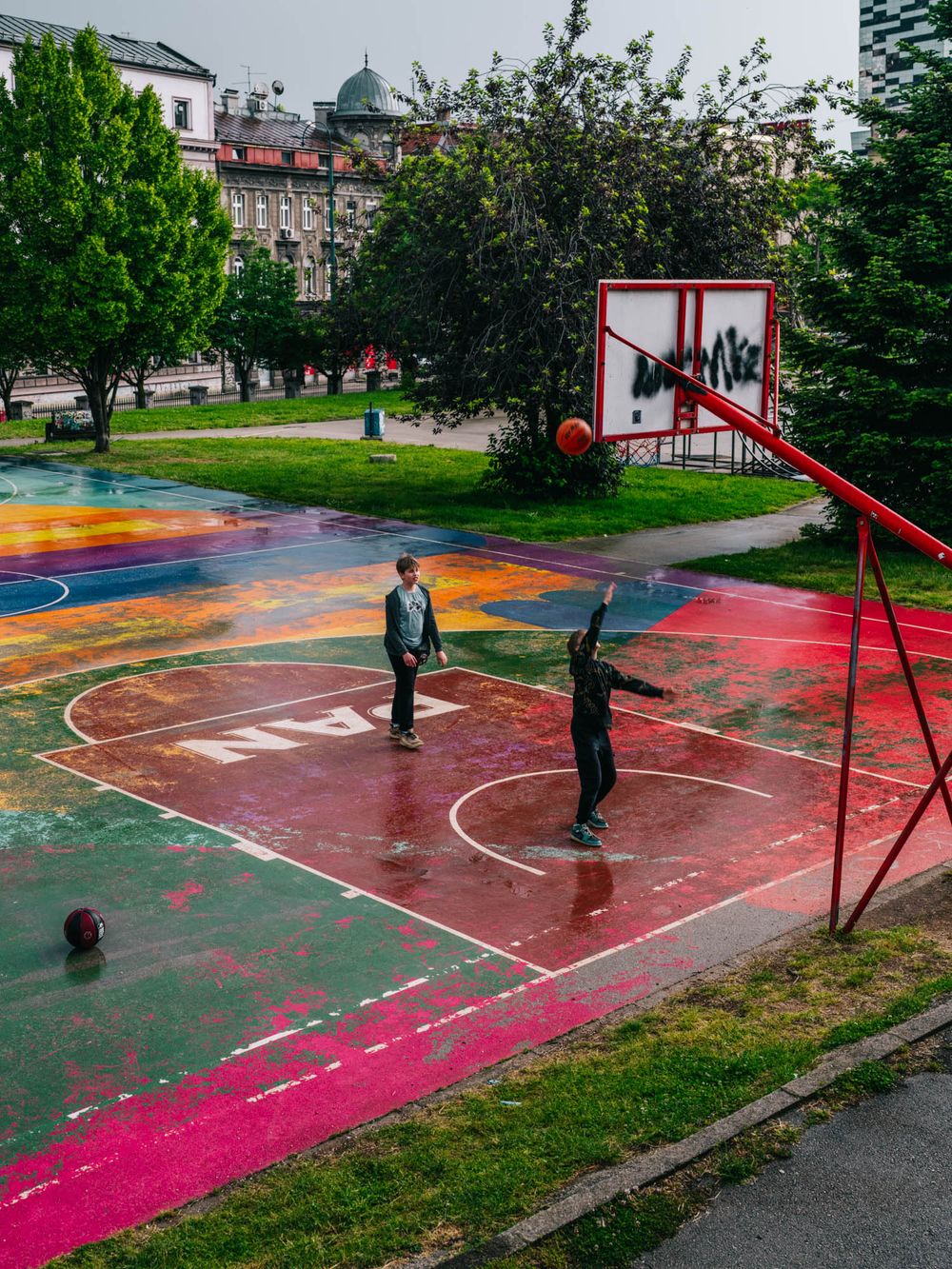 Two players shoot hoops on a vibrant, colorfully painted outdoor basketball court surrounded by lush green trees.