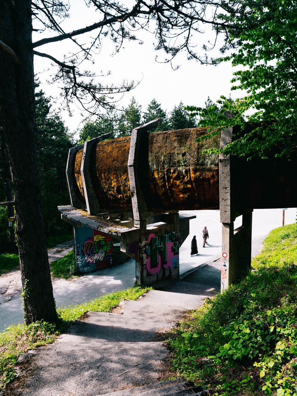 Abandoned bobsled track with graffiti-covered concrete structures surrounded by tall trees in a forested area.