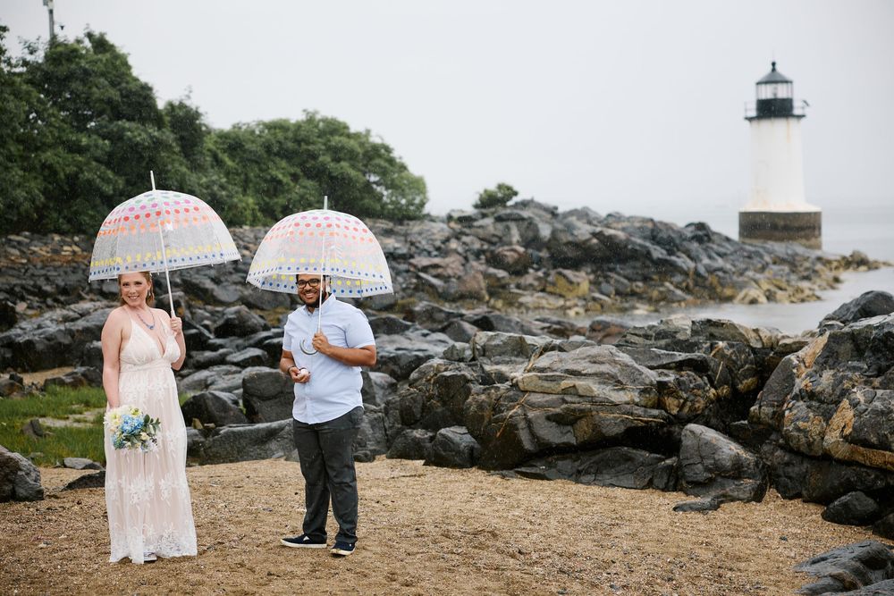 Couple holding decorative umbrellas on rocky beach near lighthouse on overcast wedding day.