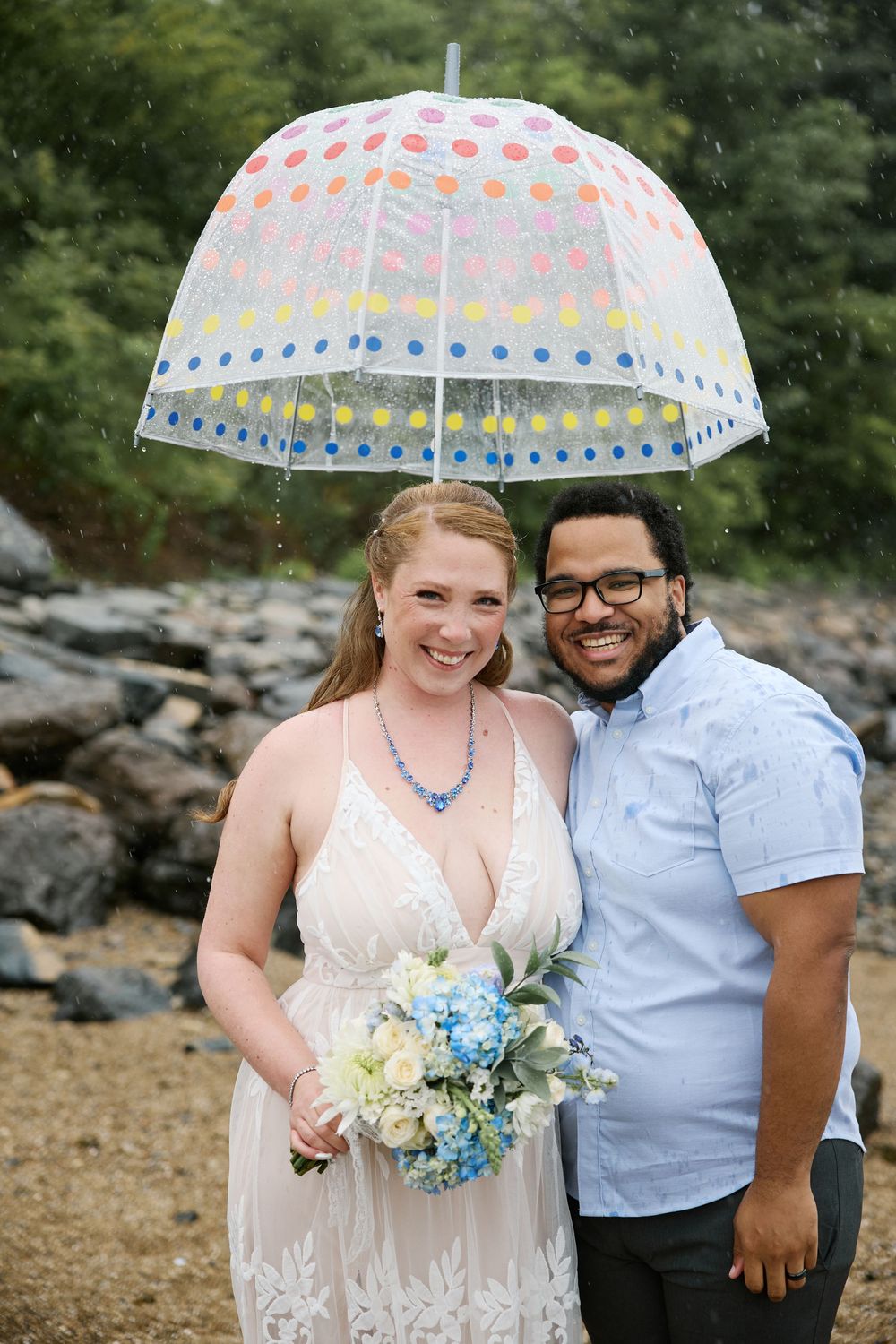Smiling newlywed couple sharing a colorful polka dot umbrella in the rain, bride holding blue and white bouquet.