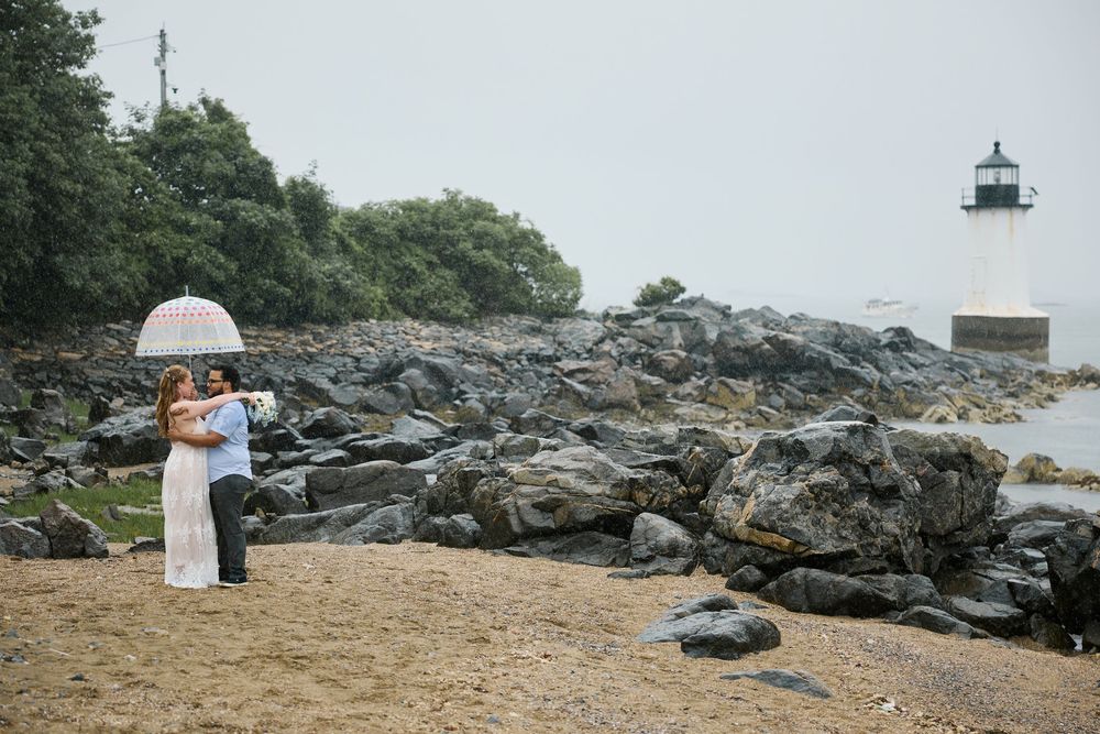 Couple embracing on rocky beach with umbrella near lighthouse on overcast day.