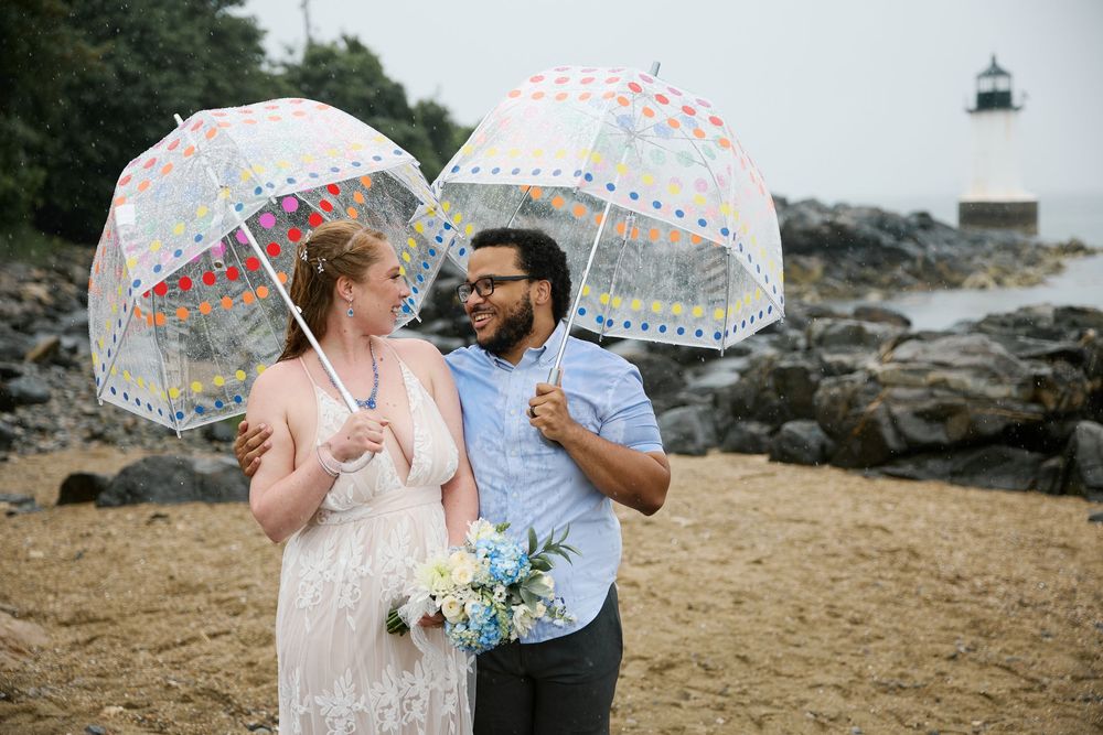 Couple holding clear polka dot umbrellas on a rocky beach near a lighthouse, bride in white dress with bouquet.