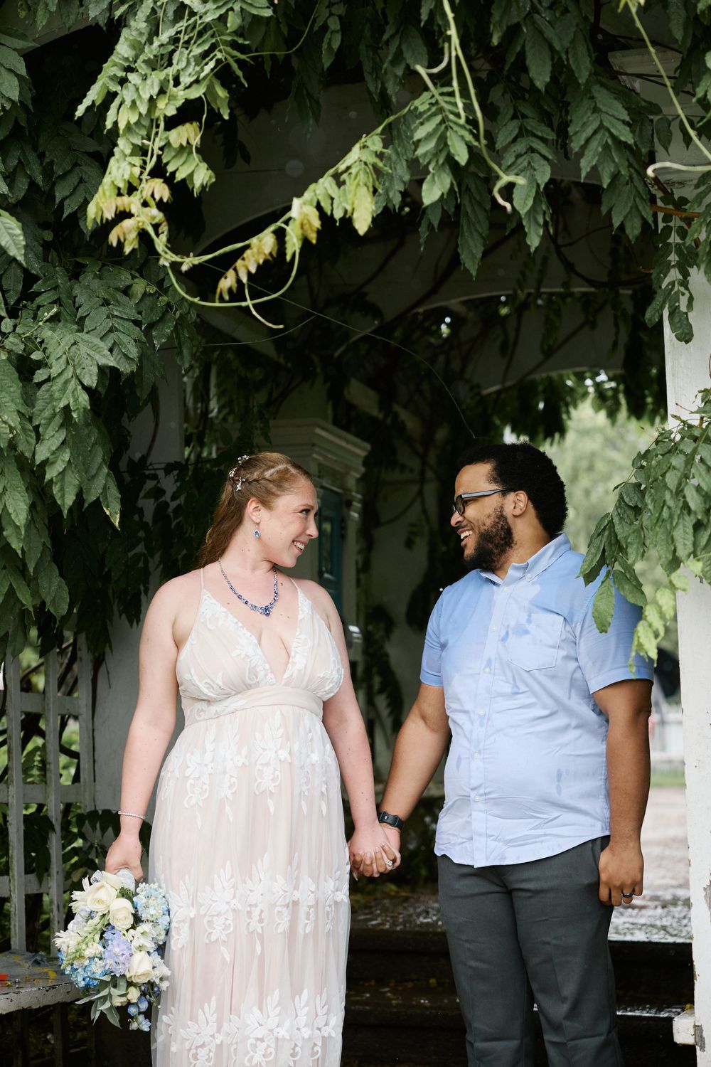 A bride and groom hold hands and smile at each other under lush green vines outdoors on their wedding day.