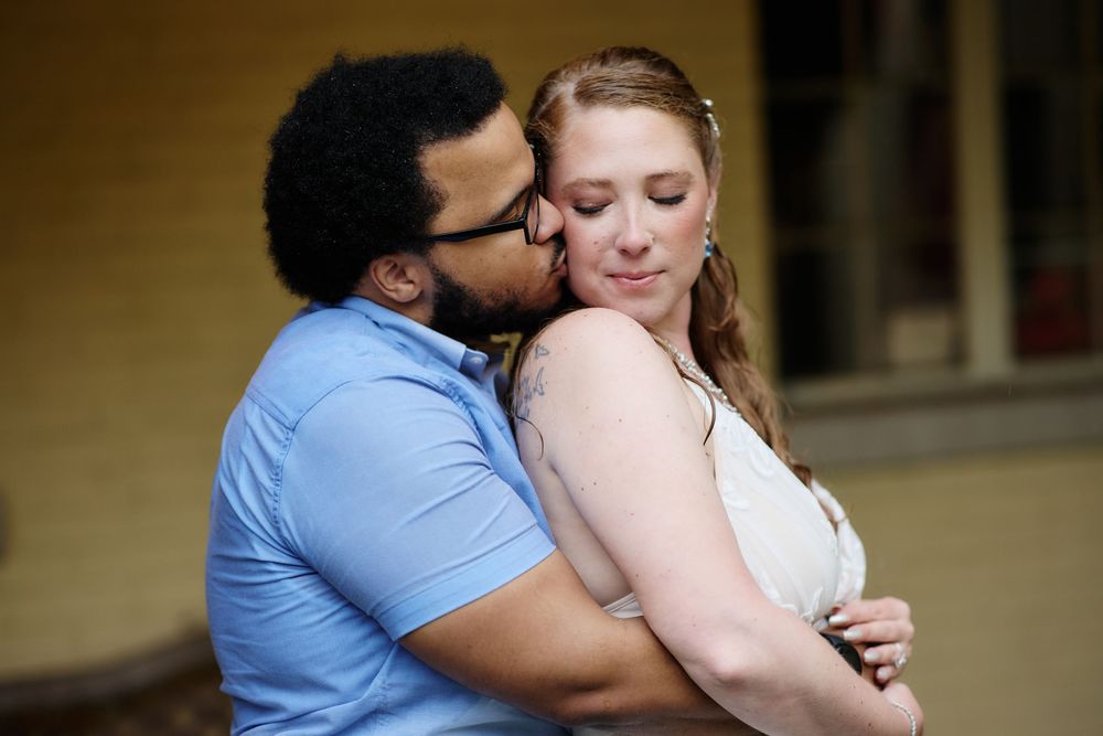A man in a blue shirt kisses a woman in a white dress on the cheek as they embrace tenderly.