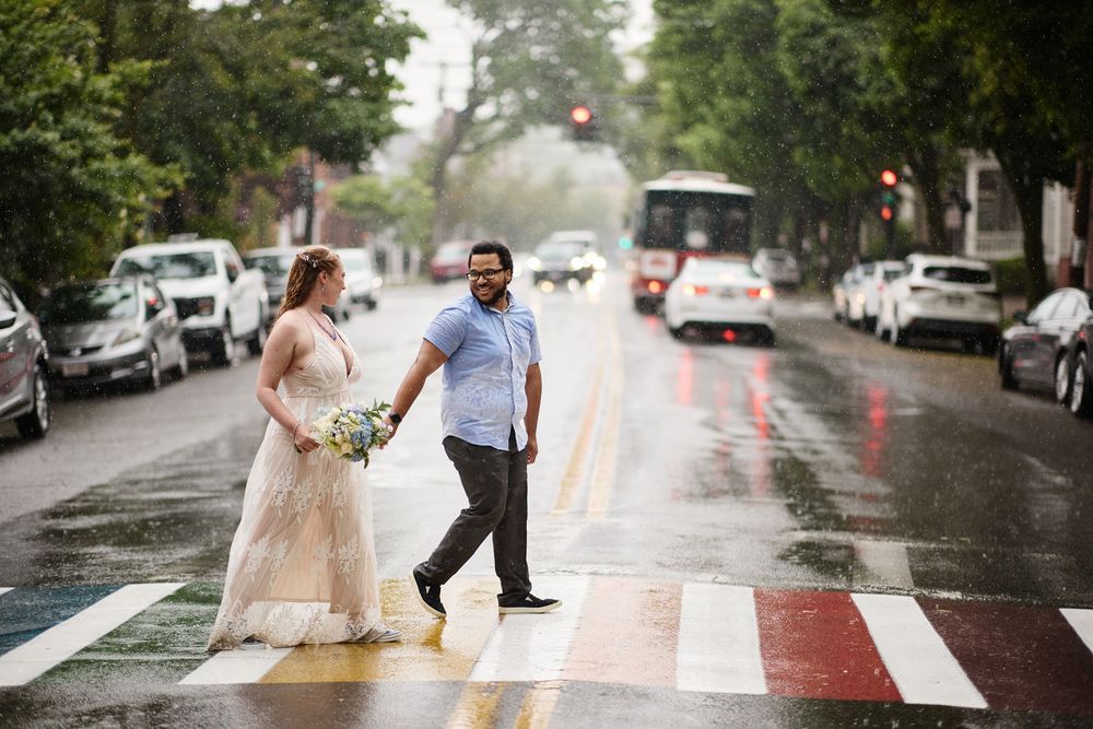 Bride and groom crossing a rainbow crosswalk in the rain on a city street, bride holding a bouquet.