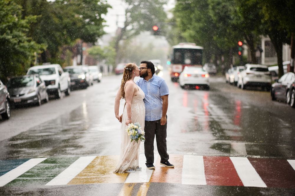 Bride and groom kissing on a rainbow crosswalk in the rain on a city street, with a bus and parked cars in the background.