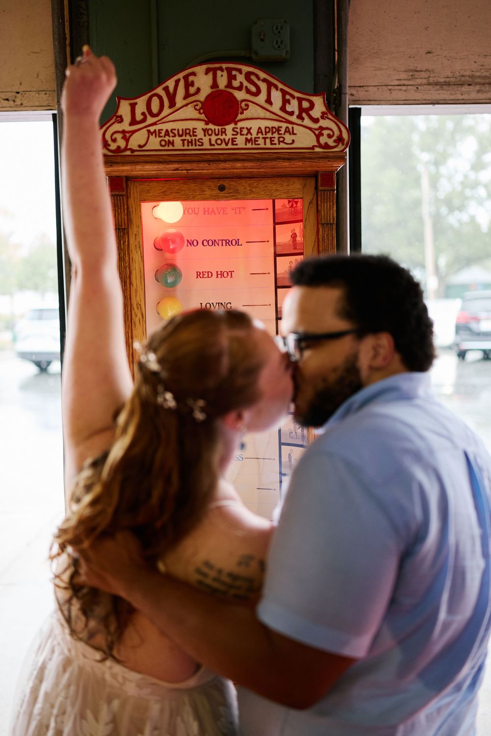 Couple kissing in front of a vintage Love Tester arcade machine, bride raising her arm in celebration.