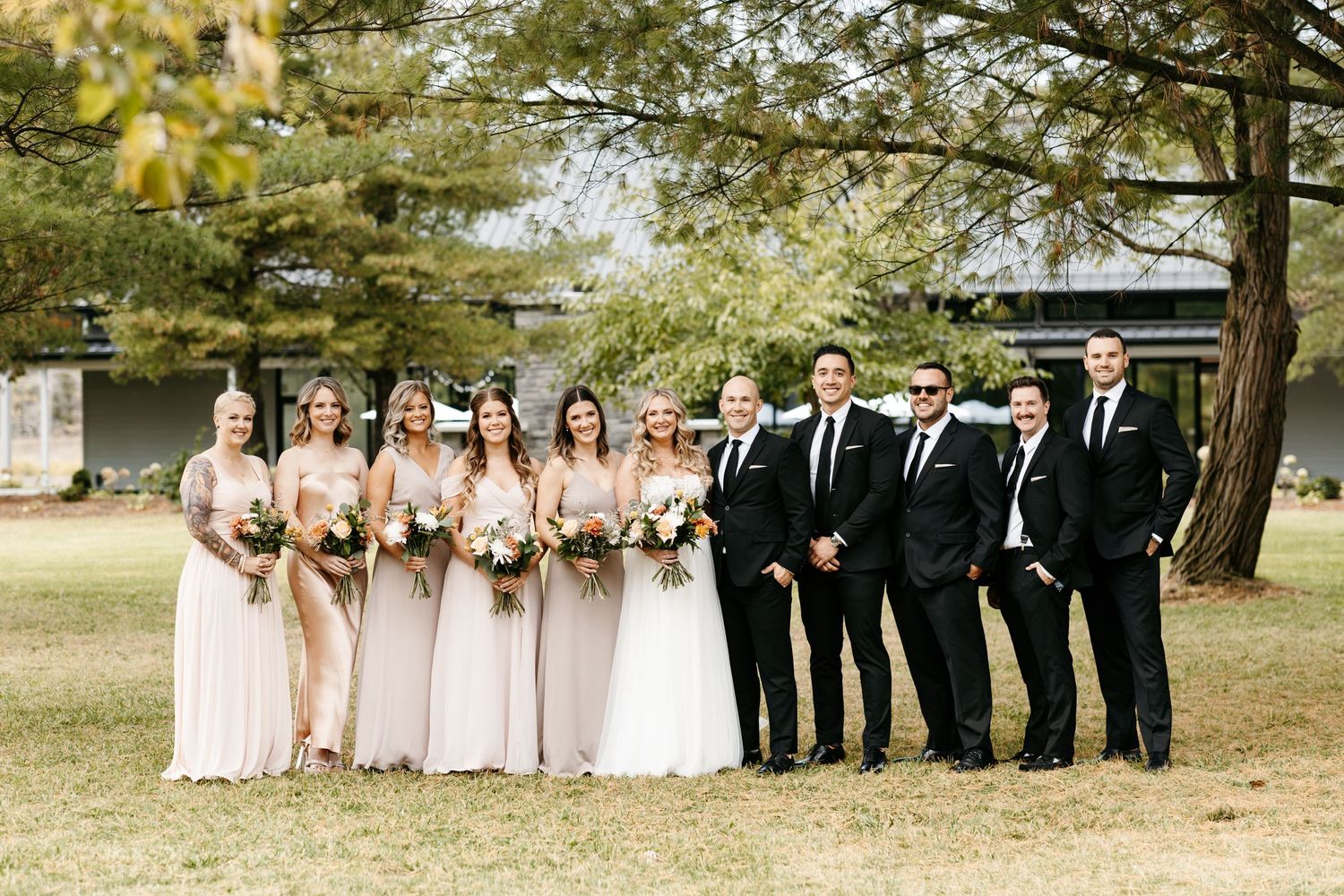 Wedding party in pink dresses and black suits posed together on lawn under trees.