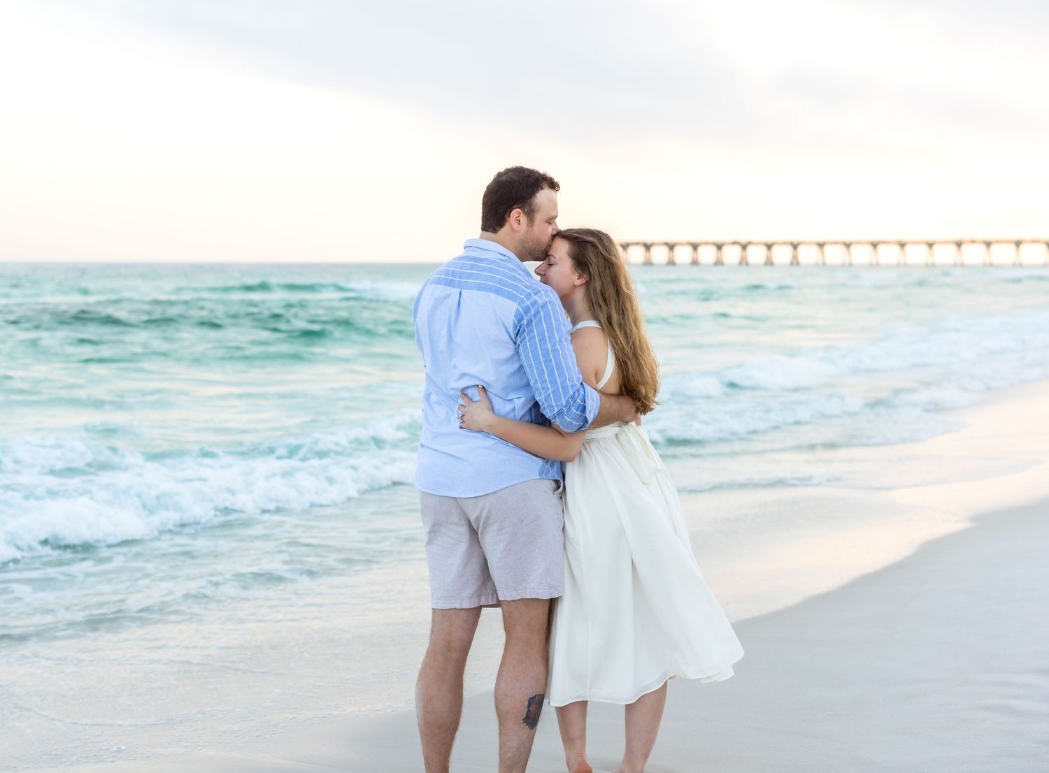 A romantic couple shares a tender moment on a white sandy beach with a wooden pier and turquoise ocean waves in view.