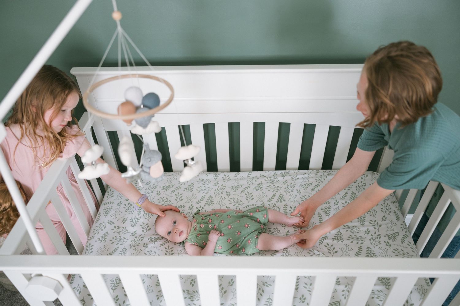Two children sitting on a porch swing holding a baby in a green romper on a sunny day.