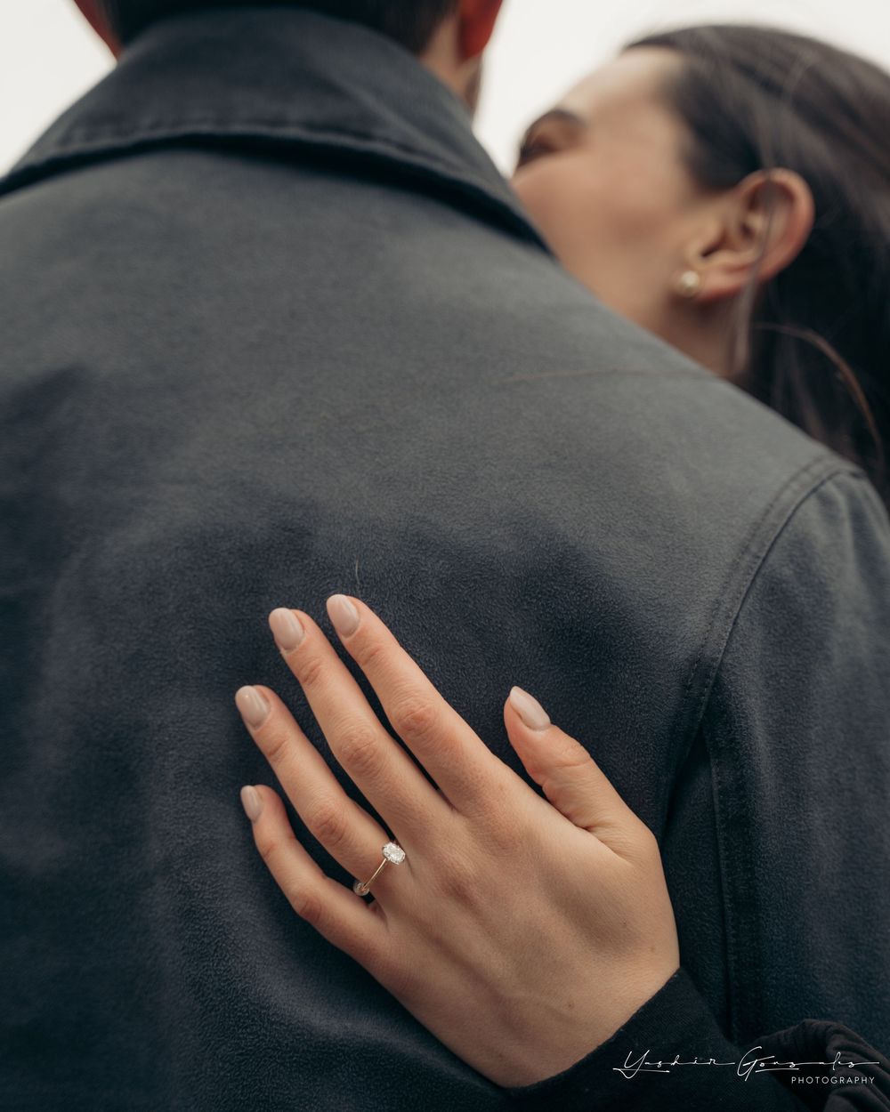 Close-up of womans hand with solitaire engagement ring resting on mans dark jacket as they embrace.

Vancouver Canada Scene