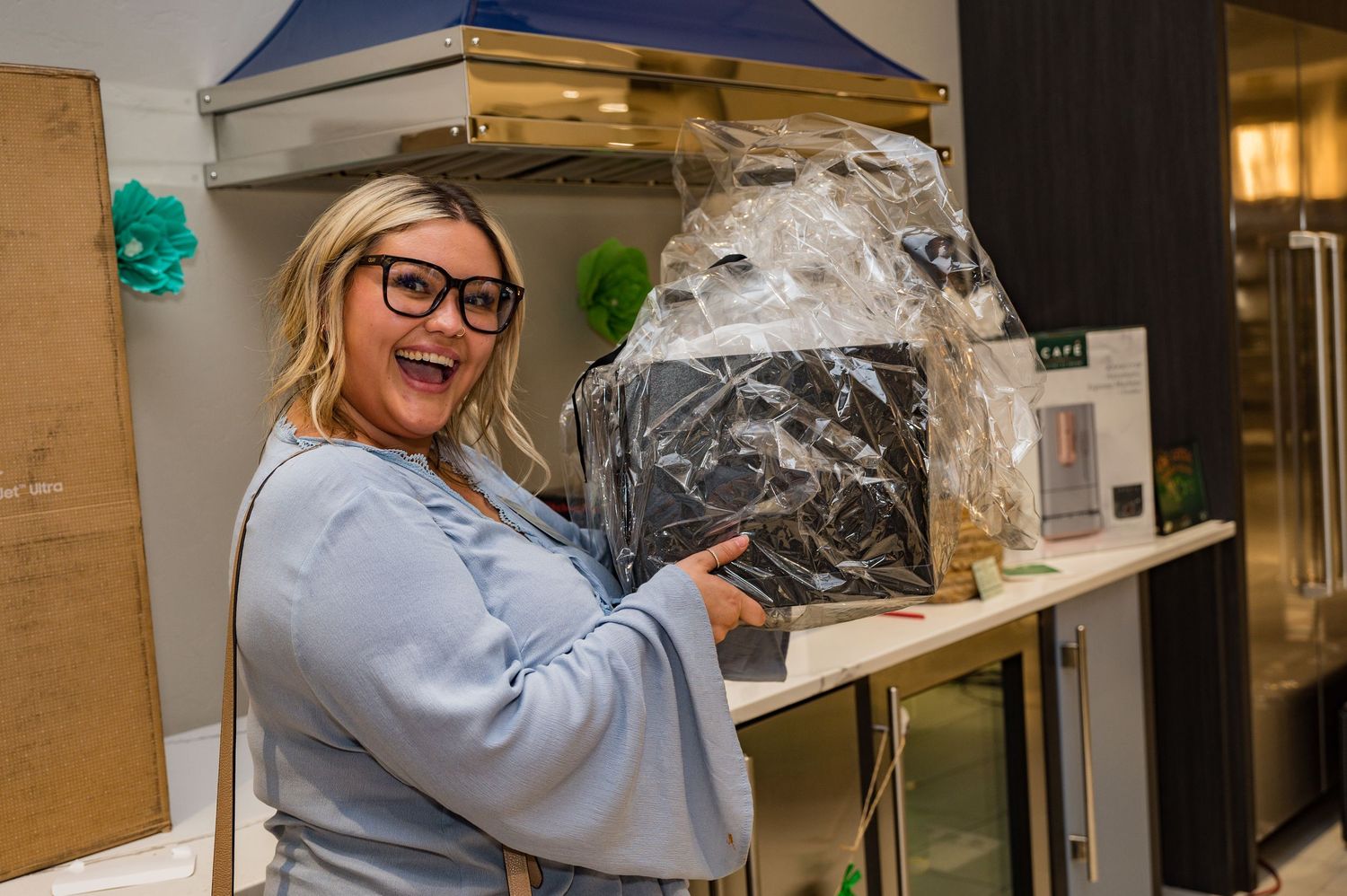 Excited blonde woman with glasses holding a wrapped appliance in a modern kitchen showroom.