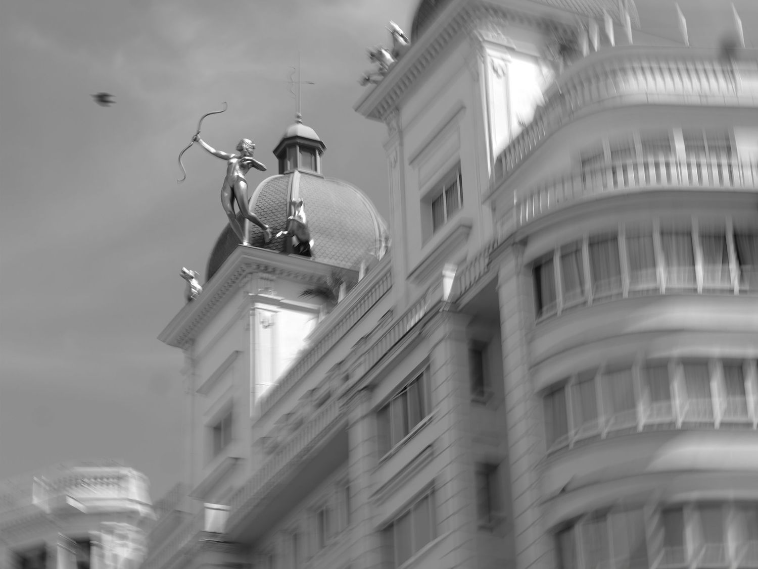 Black and white photo of a classical archer statue atop a domed building with ornate architecture in the background.