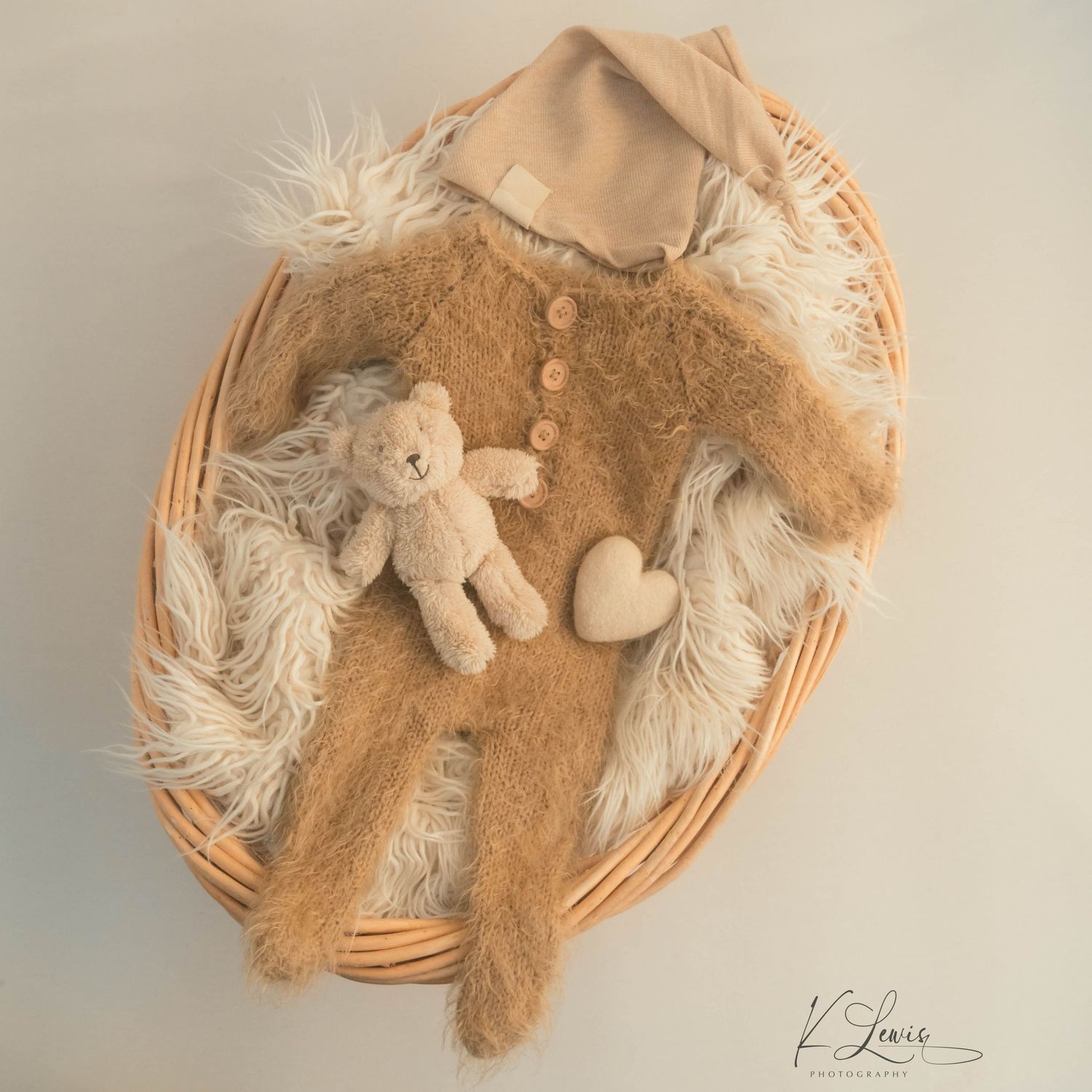 Brown knit baby romper with matching hat and teddy bear arranged in a wicker basket with fur lining.