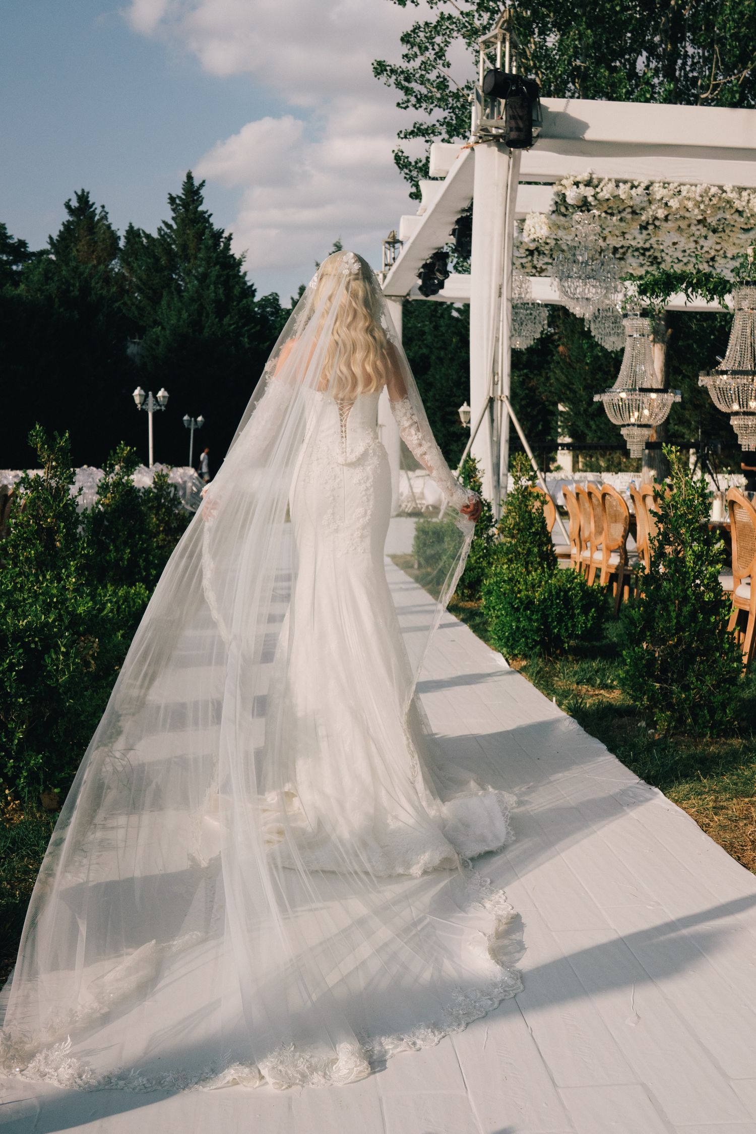 A flowing white wedding dress with a long veil cascades across a white platform in an outdoor garden setting.