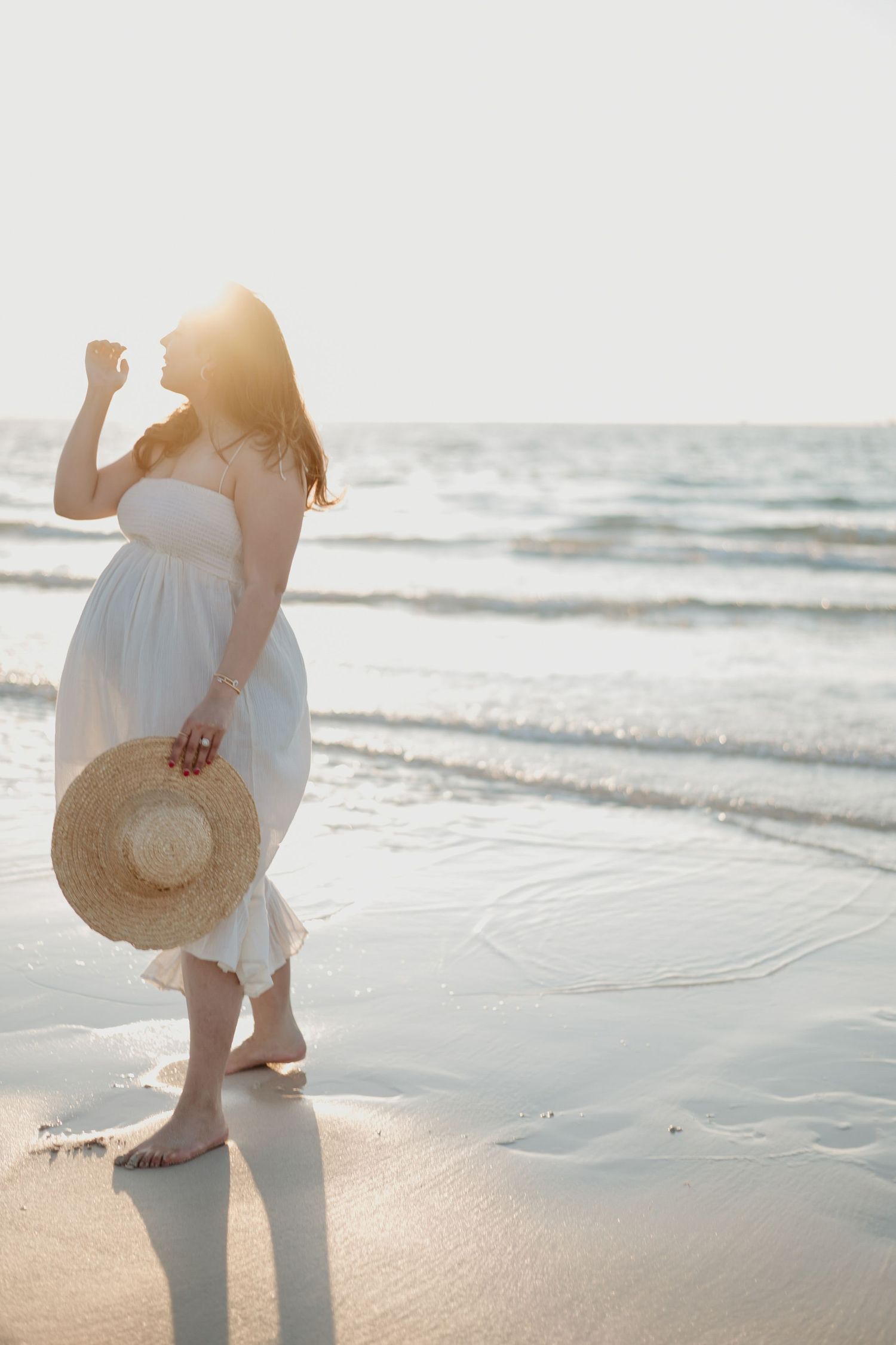 Pregnant woman in white dress standing on beach at sunset holding straw hat.