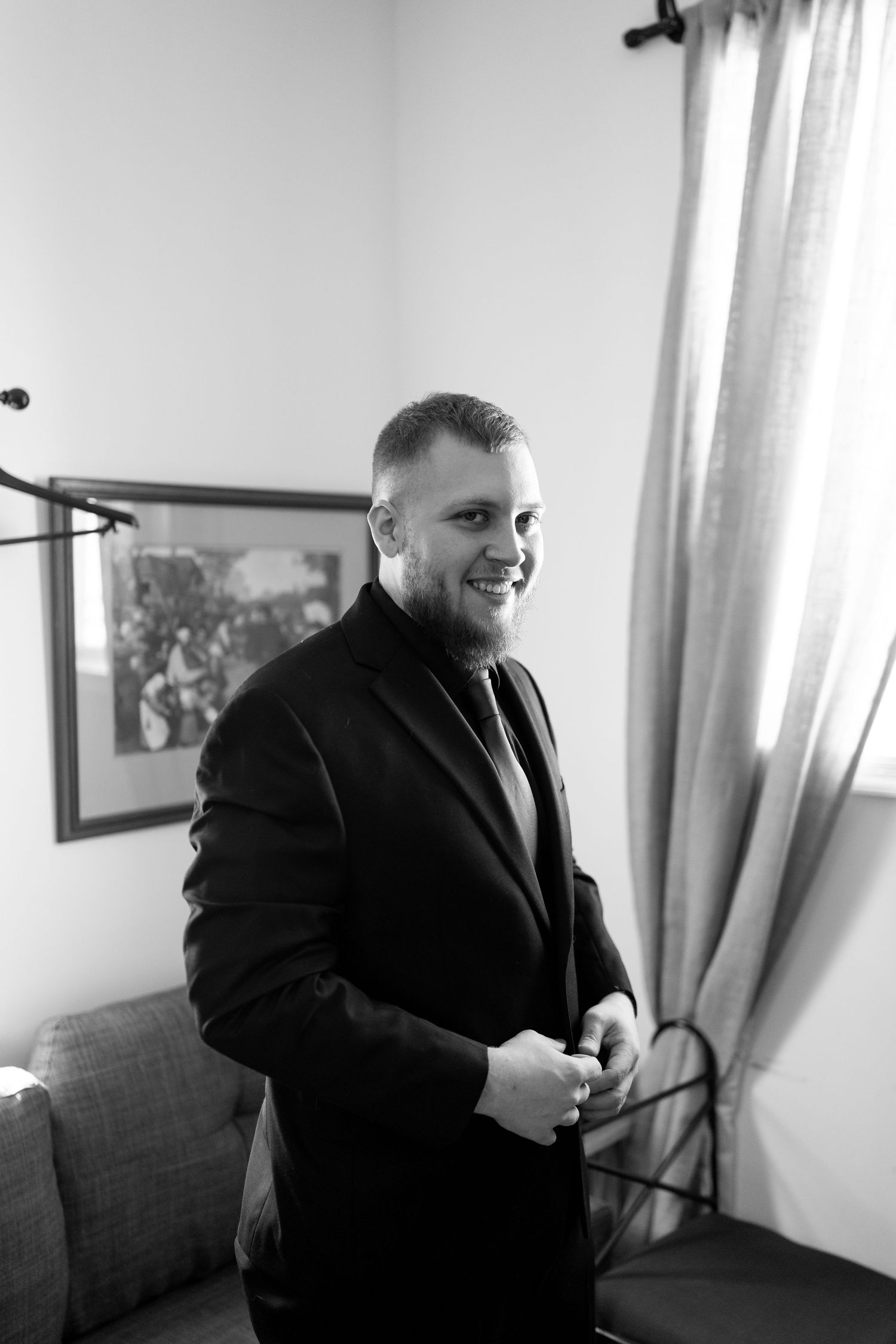 Black and white portrait of smiling groom buttoning his suit jacket before wedding ceremony.
