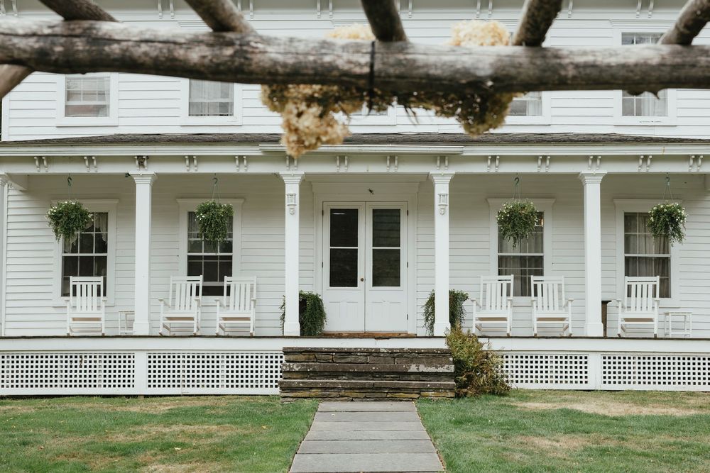 Classic white farmhouse with a wide covered porch, columns, and hanging plants along the front facade.