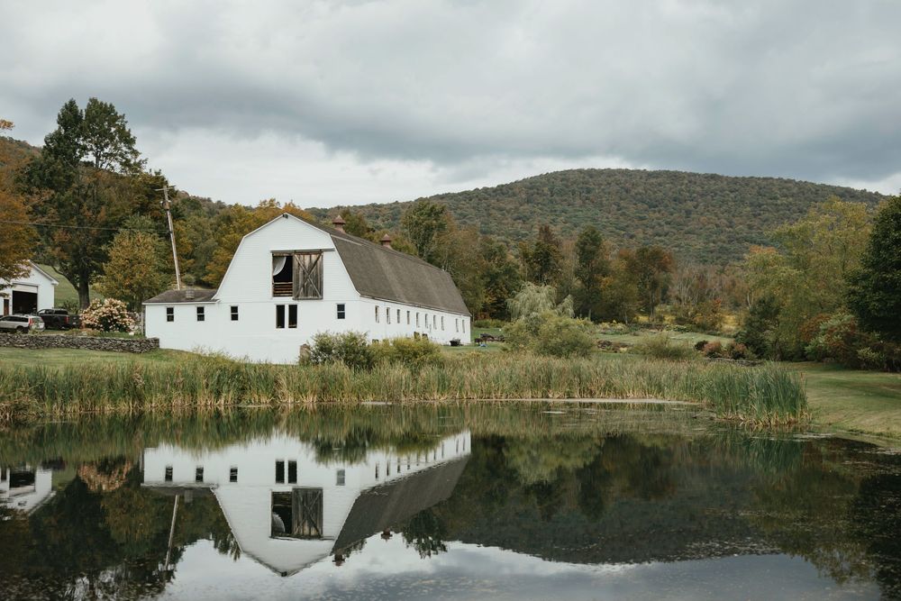 A serene white barn and pond reflect in still water against rolling hills and autumn foliage in Vermont.