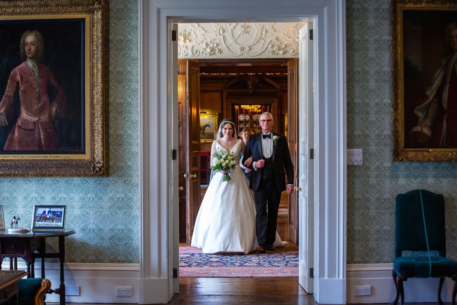 Bride walking through ornate doorway with escort in a grand stately home with portrait paintings on walls.