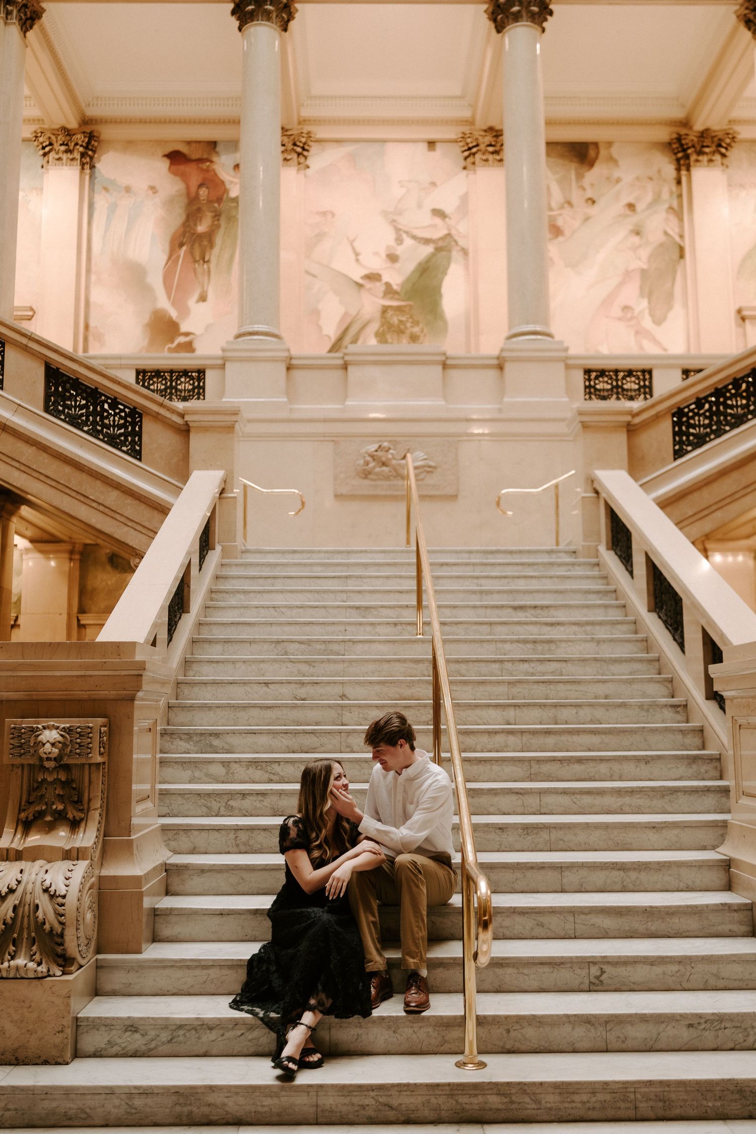 Couple sitting close together on grand marble staircase beneath ornate murals in historic building.