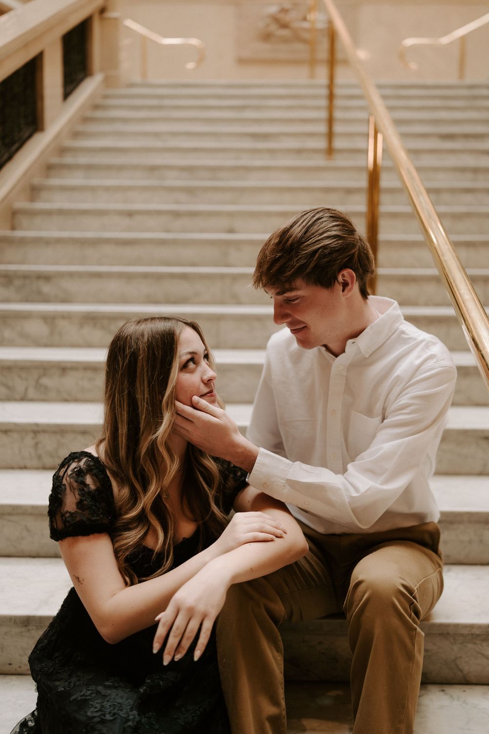 Couple sitting on marble stairs, man tenderly touching woman's face during romantic engagement photo session.