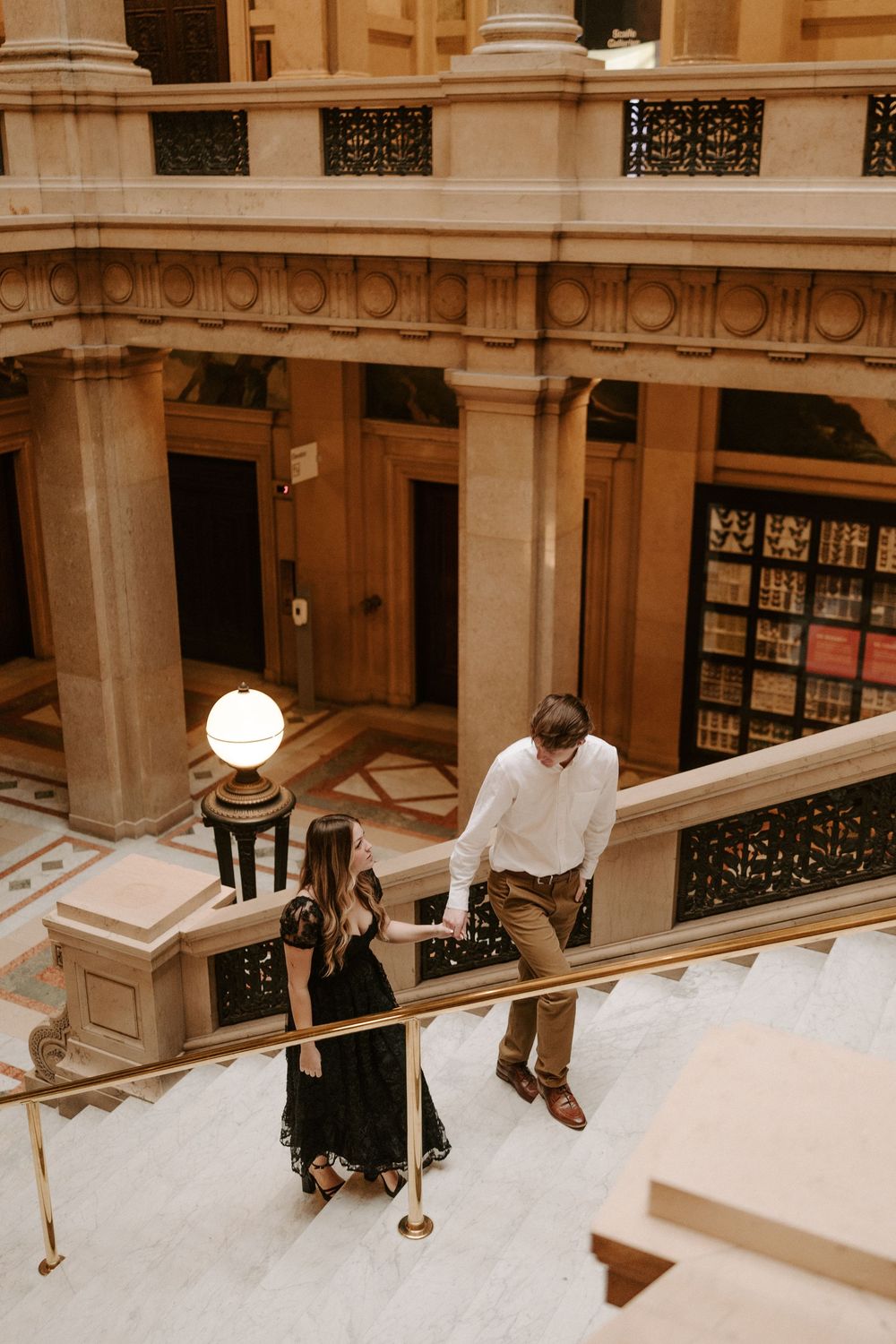 Couple holding hands walking up grand marble staircase in elegant historic building interior.
