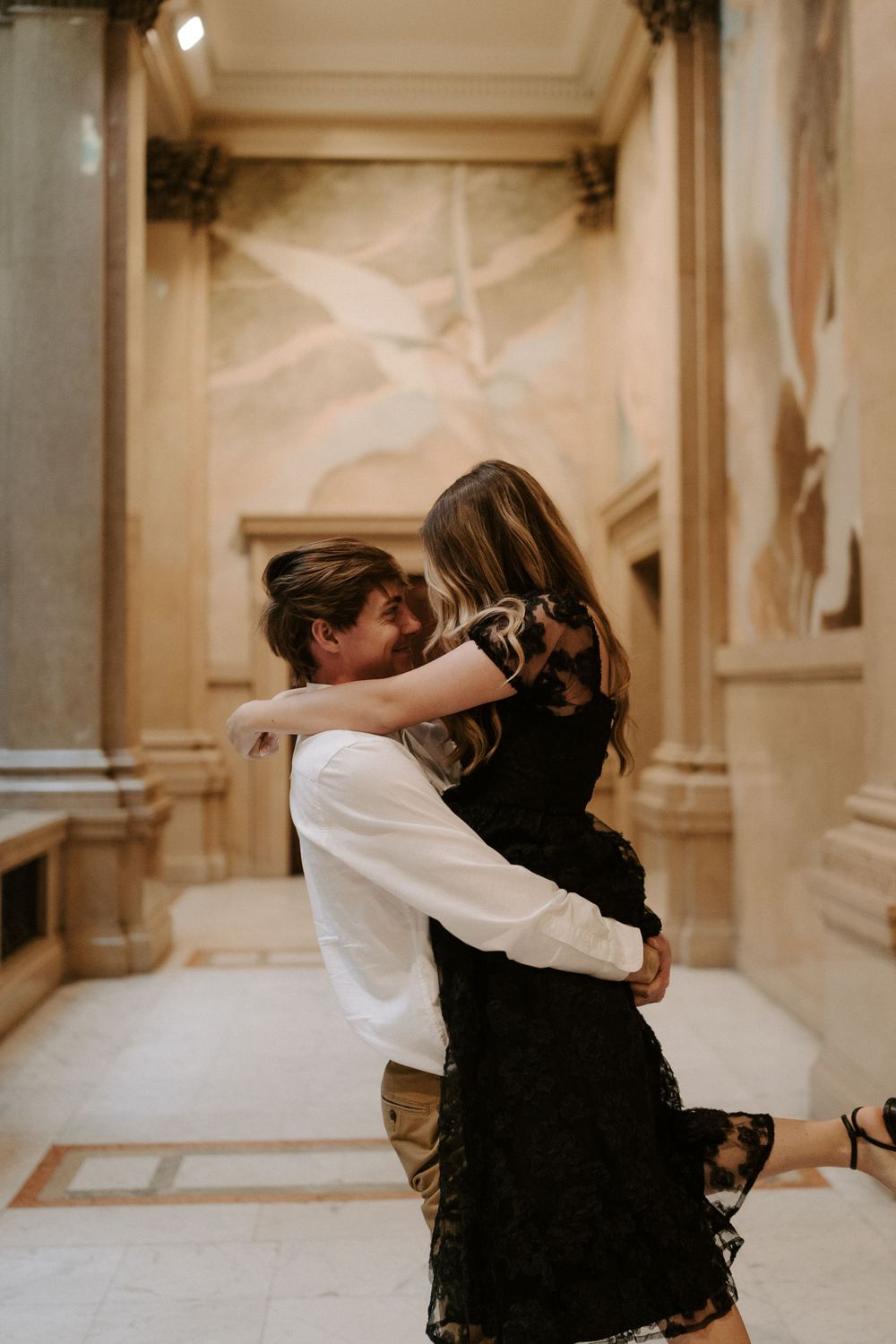 Couple sharing joyful embrace in ornate historic building with decorative murals and marble columns.