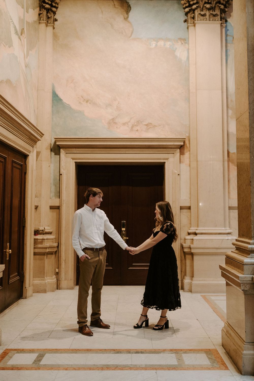 Couple holding hands facing each other in elegant hall with classical murals and marble columns.