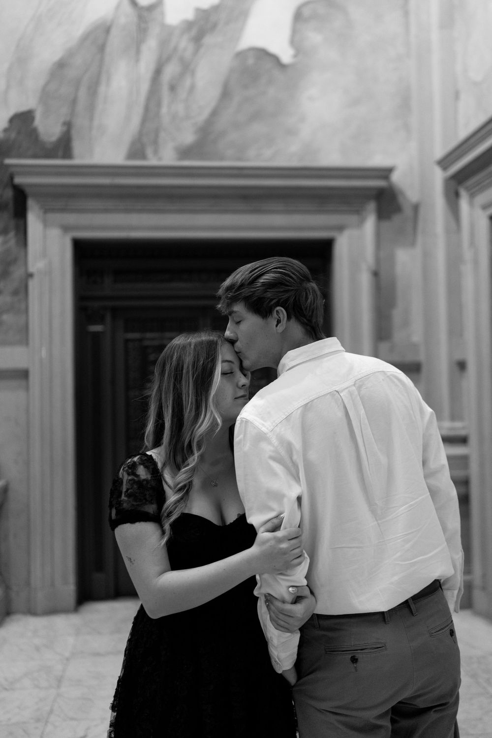 Black and white photo of couple sharing a kiss in elegant hall with classical architecture.