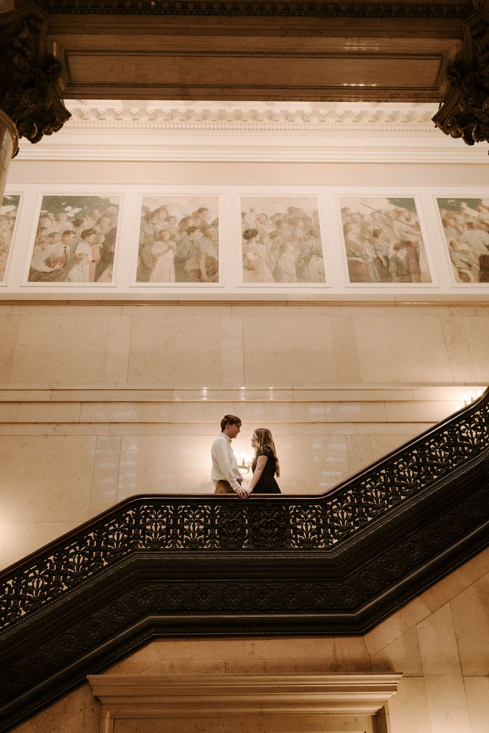 Couple standing close on ornate iron staircase beneath classical murals in grand historic building.